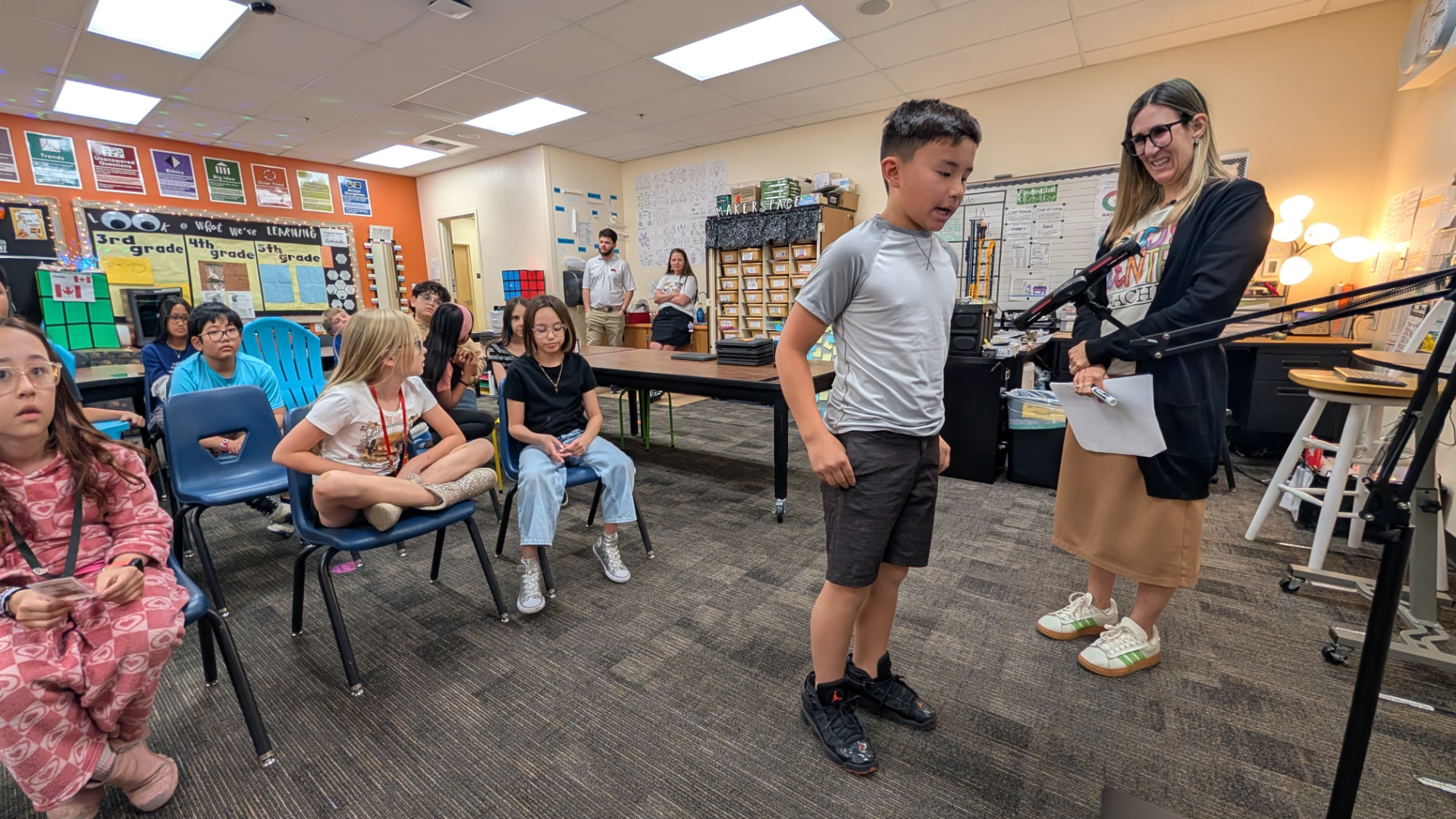 A student speaking at the front of a classroom while a teacher watches nearby.